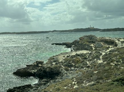 A scenic coastal view with rocky shores, turquoise water, and a distant lighthouse under a cloudy sky. Overlaid text reads…