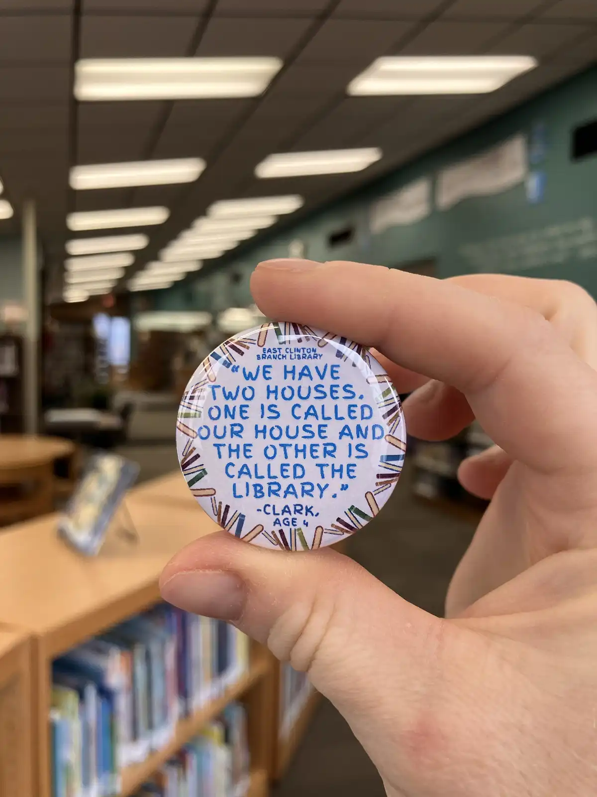 A hand holds a circular button in a library, featuring a child's quote about loving the library so much it's like a second…