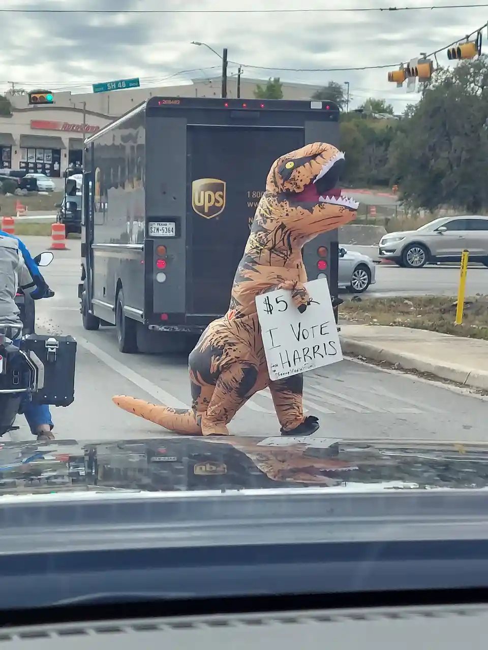 A person in a full-body inflatable t-rex costume stands on a street corner holding a handwritten sign that reads '$5 I VOTE…
