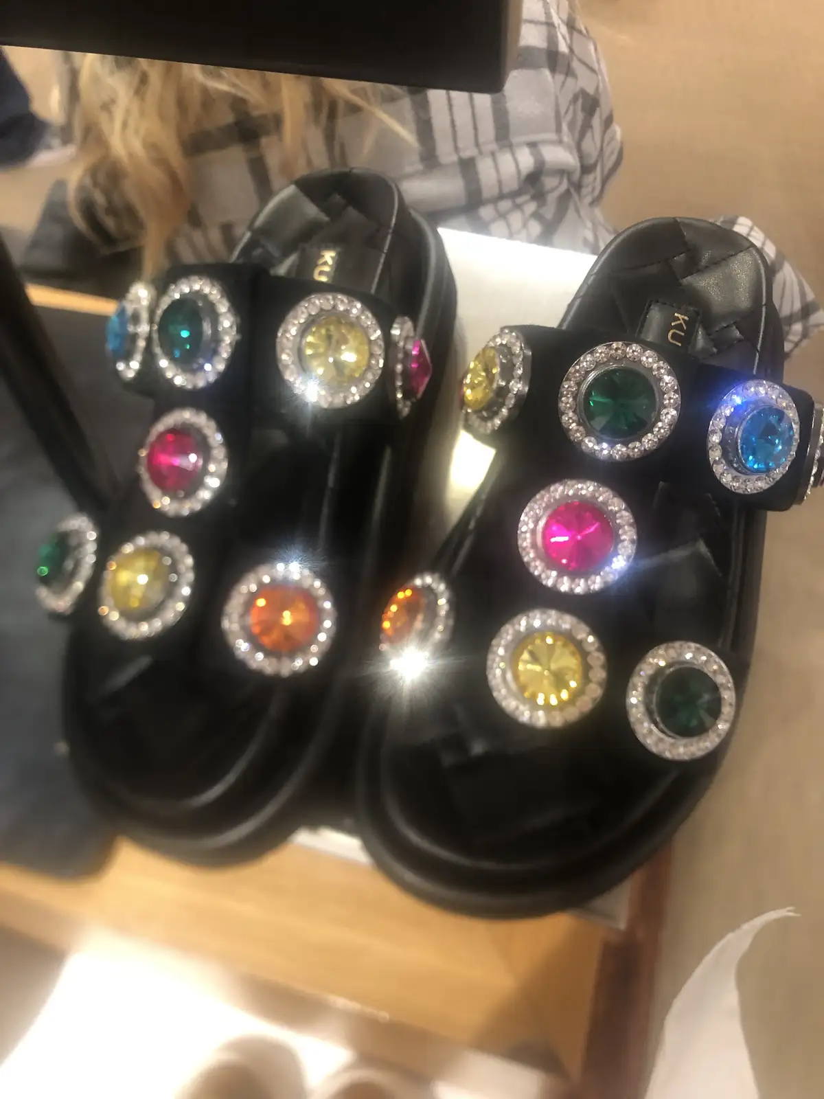 A close-up photo shows a pair of black, quilted, open-toed sandals heavily adorned with numerous large, colorful gemstones…