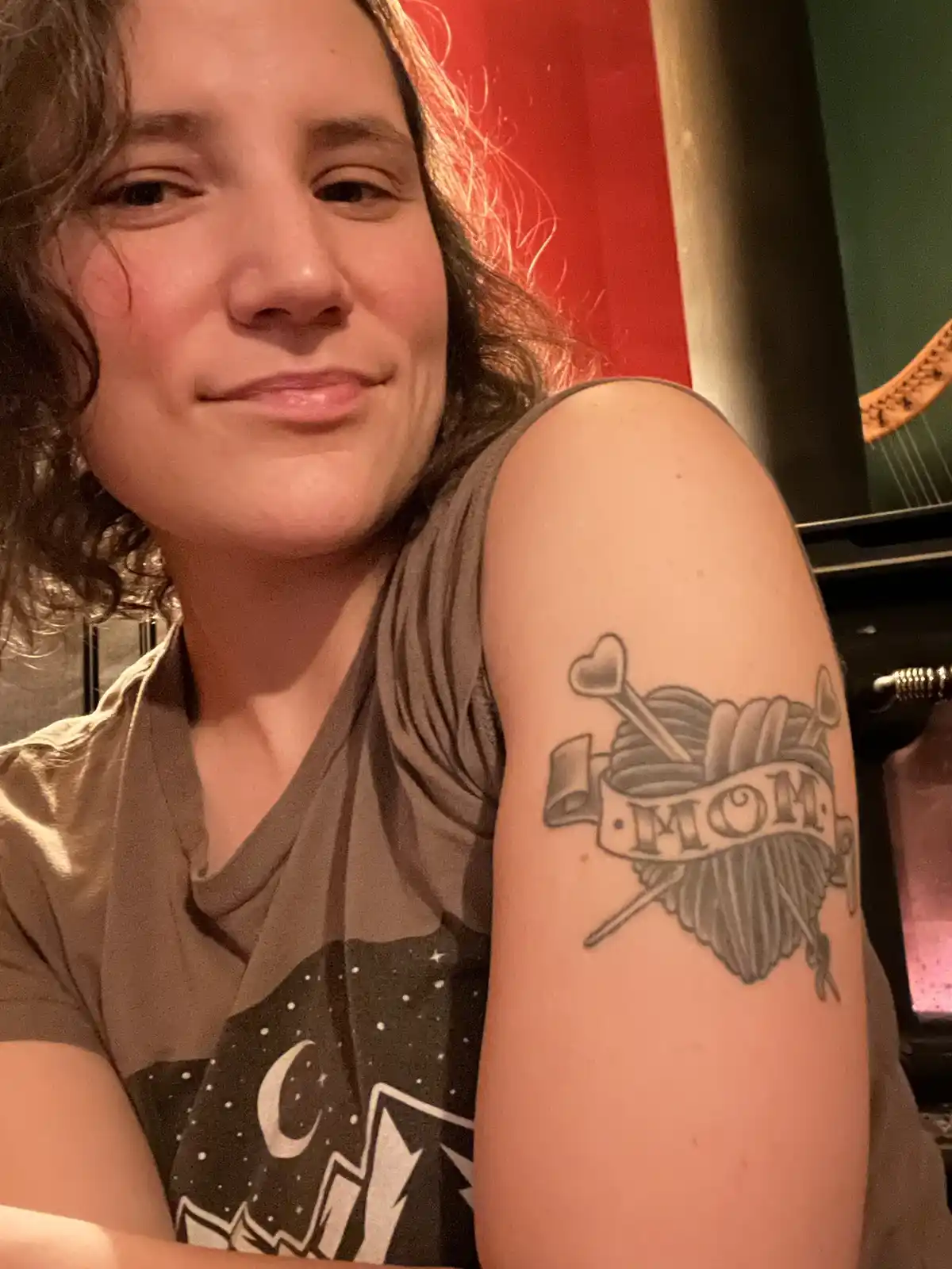 A person with dark curly hair and a brown t-shirt smiles while showing off a tattoo on their upper arm. The tattoo is a…
