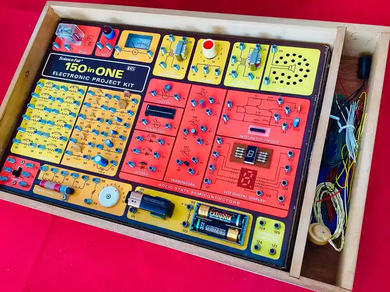 A vintage 'Science Fair 150 in ONE Electronic Project Kit' is shown in a wooden box, featuring colorful sections labeled…