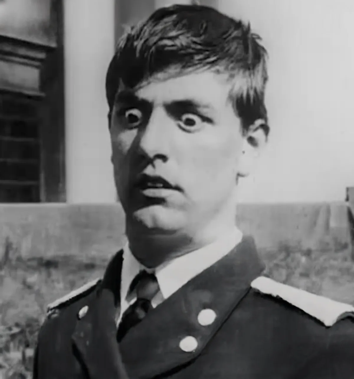 A black-and-white photo shows a young man in a military-style uniform with wide, startled eyes and a slightly open mouth…