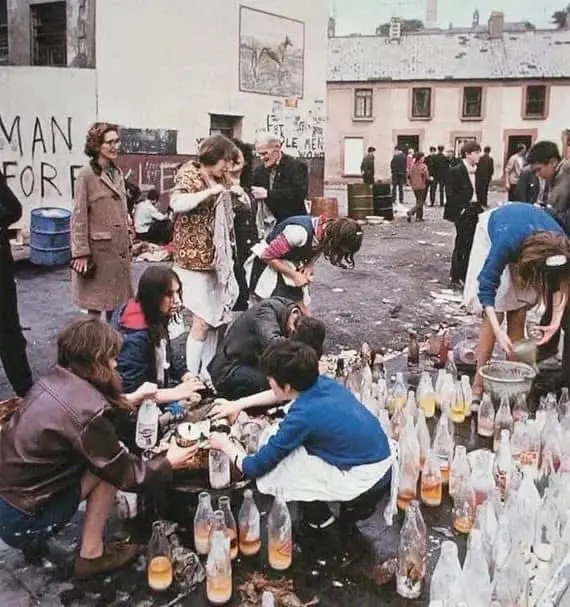 A vintage photo shows a group of people, mostly young adults, gathered around a table outdoors in a gritty urban courtyard…