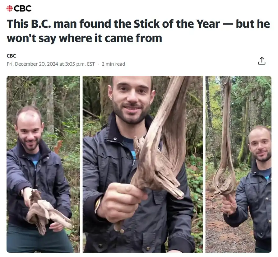 A man in a dark jacket stands in a forest holding up an unusually shaped, long, twisted piece of driftwood or tree branch…