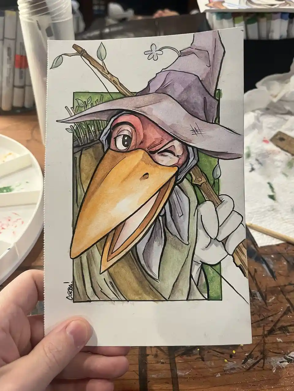 A hand holds up a watercolor drawing of a cartoonish bird with a large orange beak, wearing a purple wizard's hat adorned…
