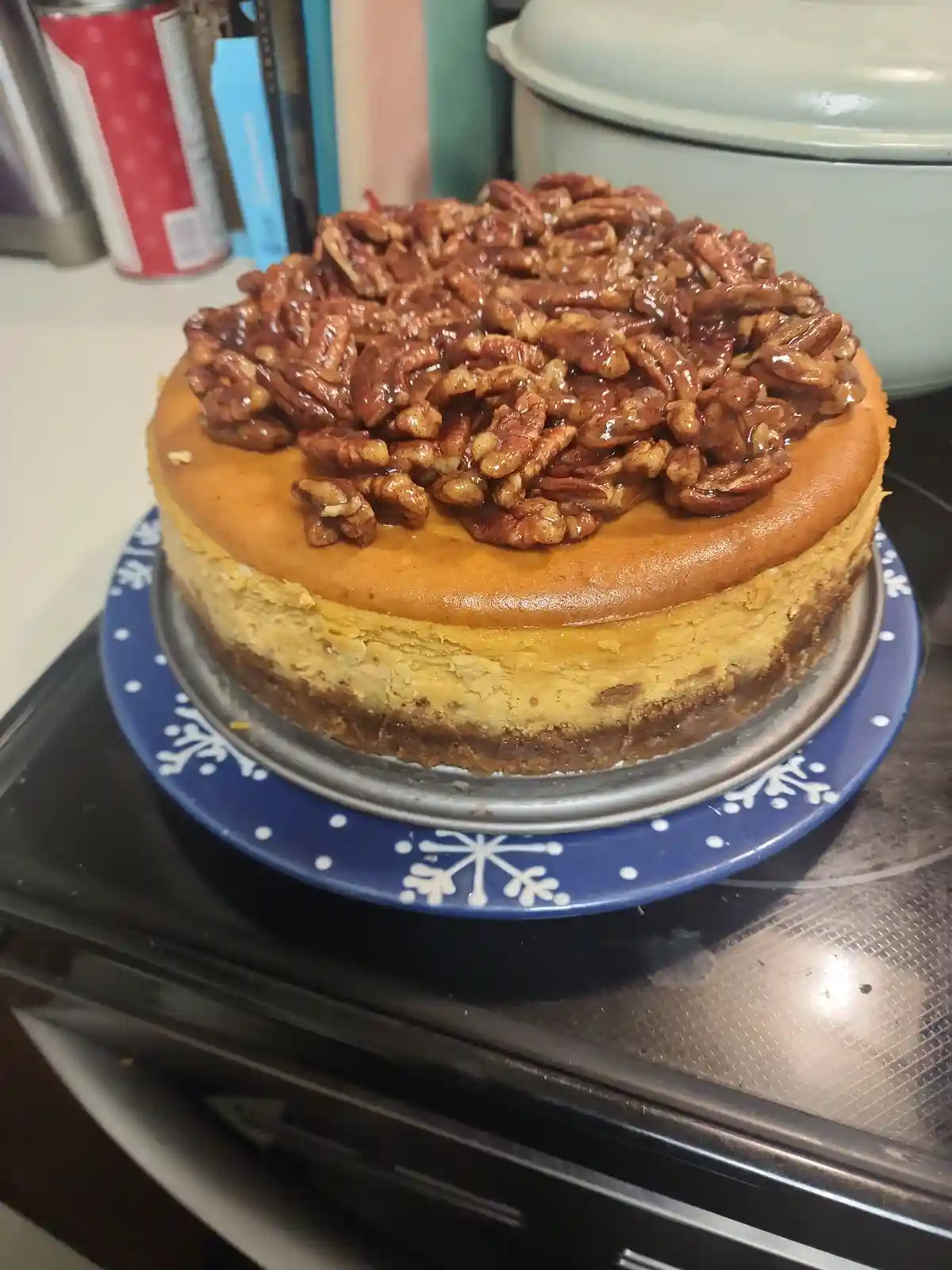 A freshly baked cheesecake with a golden-brown top and a crust, generously covered with glossy, caramelized pecans, sits on…