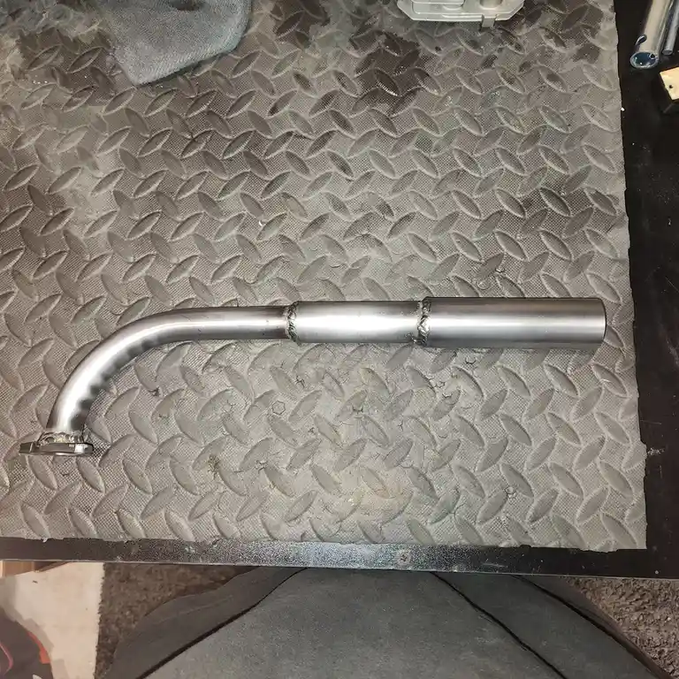 A shiny, custom-made metal exhaust pipe with welded joints lies on a diamond plate work surface. The overlaid text reads 'i…