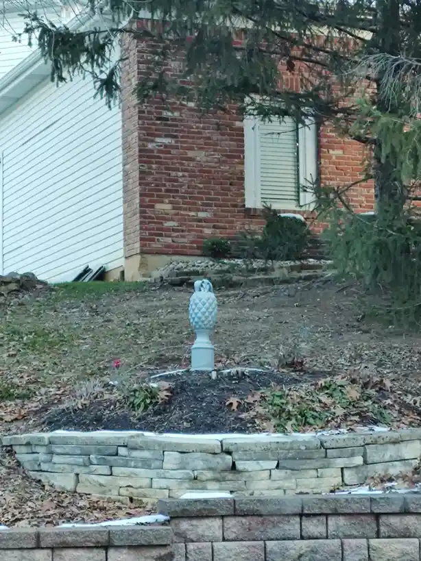 A light blue, pineapple-shaped garden ornament stands in a mulched flower bed surrounded by stone retaining walls, with a…