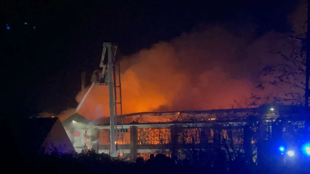 A nighttime photo shows a large building engulfed in flames and thick orange smoke, with a fire truck's ladder extended and…