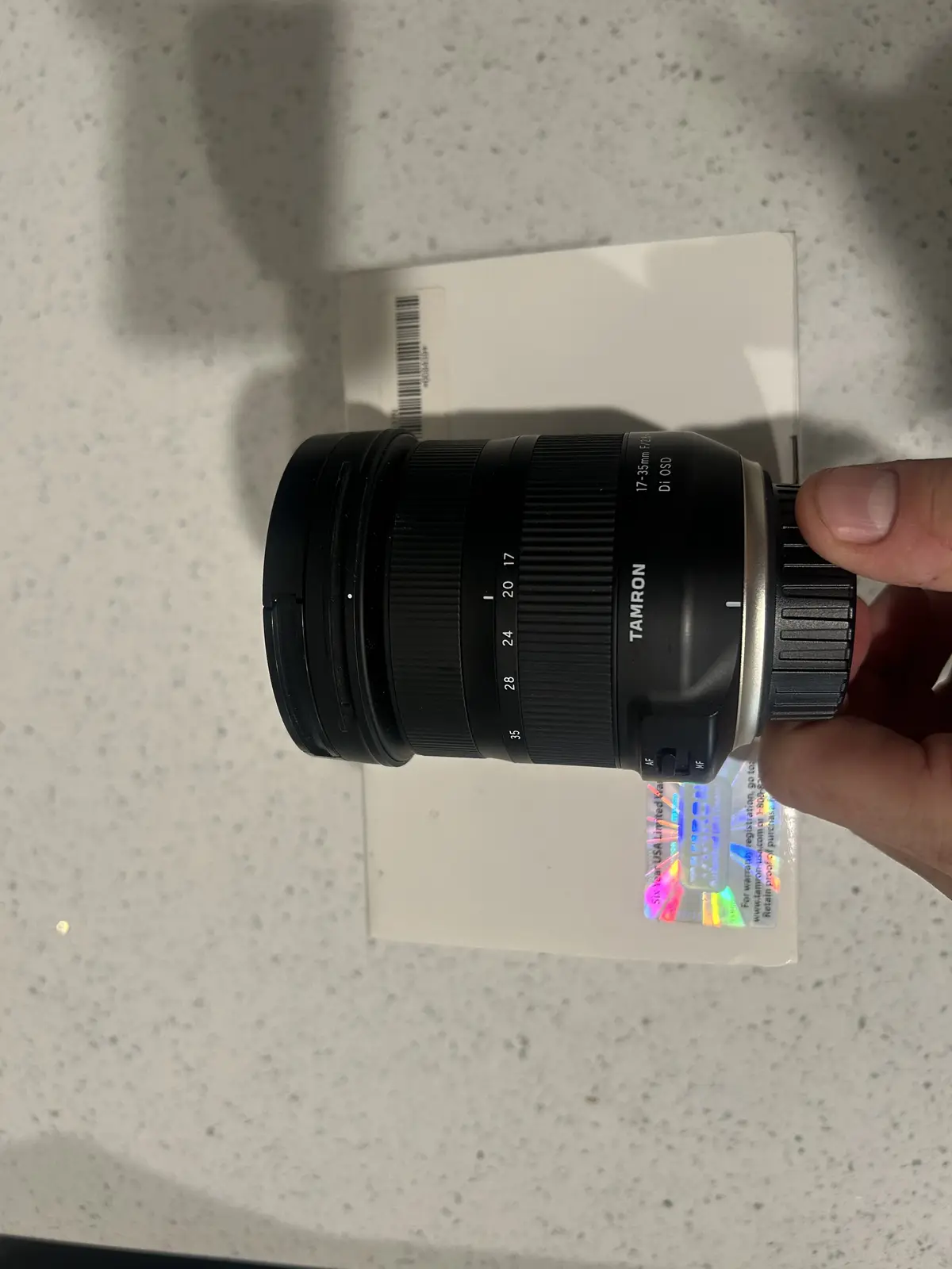 A hand holds a black Tamron 17-35mm f/2.8 Di OSD camera lens, showing its zoom ring and AF/MF switch, resting on a white box…