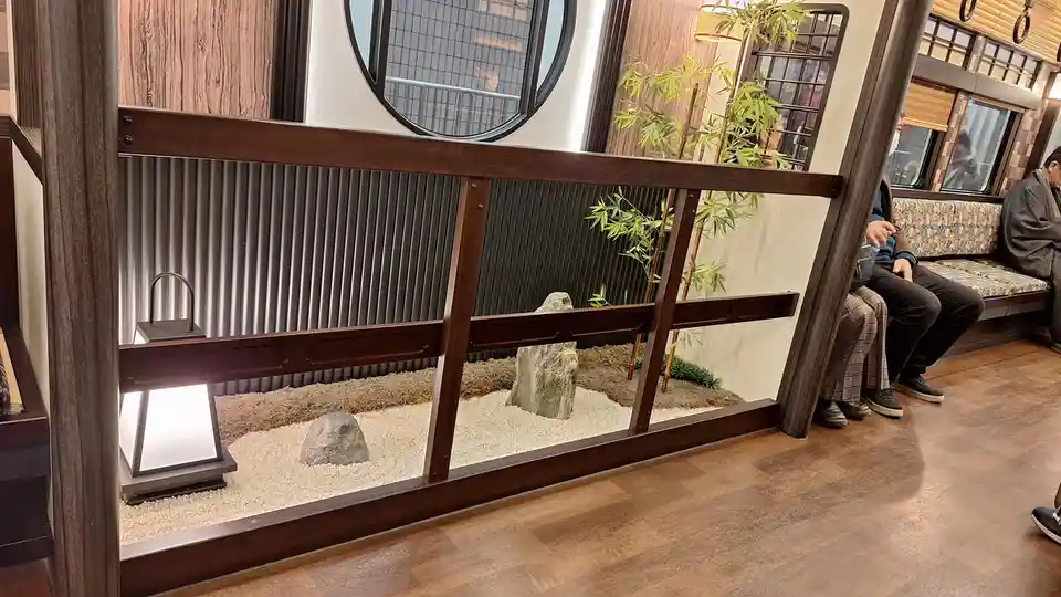 a small indoor zen garden with white gravel, two rocks, and a bamboo plant, enclosed by a dark wooden railing, with people…