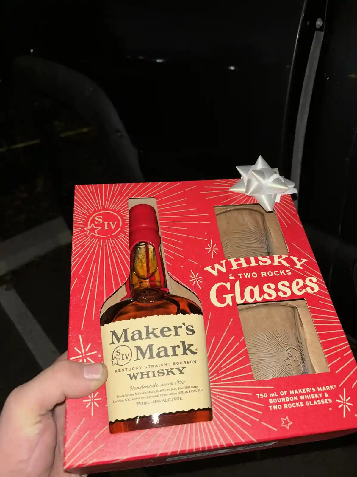 A hand holds a red gift box containing a bottle of Maker's Mark bourbon whisky and two rocks glasses, topped with a silver…
