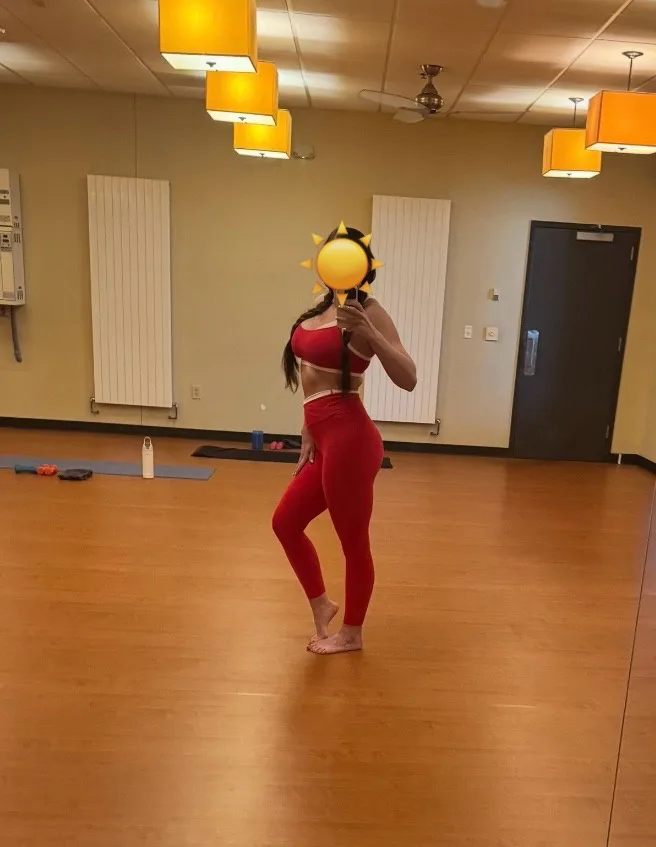 A woman in a red sports bra and leggings stands barefoot in a gym or yoga studio, holding a sun-shaped object over her face…