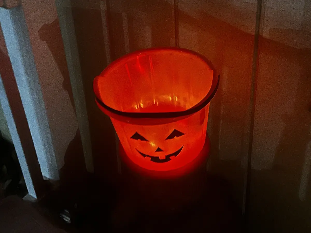 A glowing orange plastic bucket with a carved jack-o'-lantern face sits in a dimly lit room, casting a warm red light. The…
