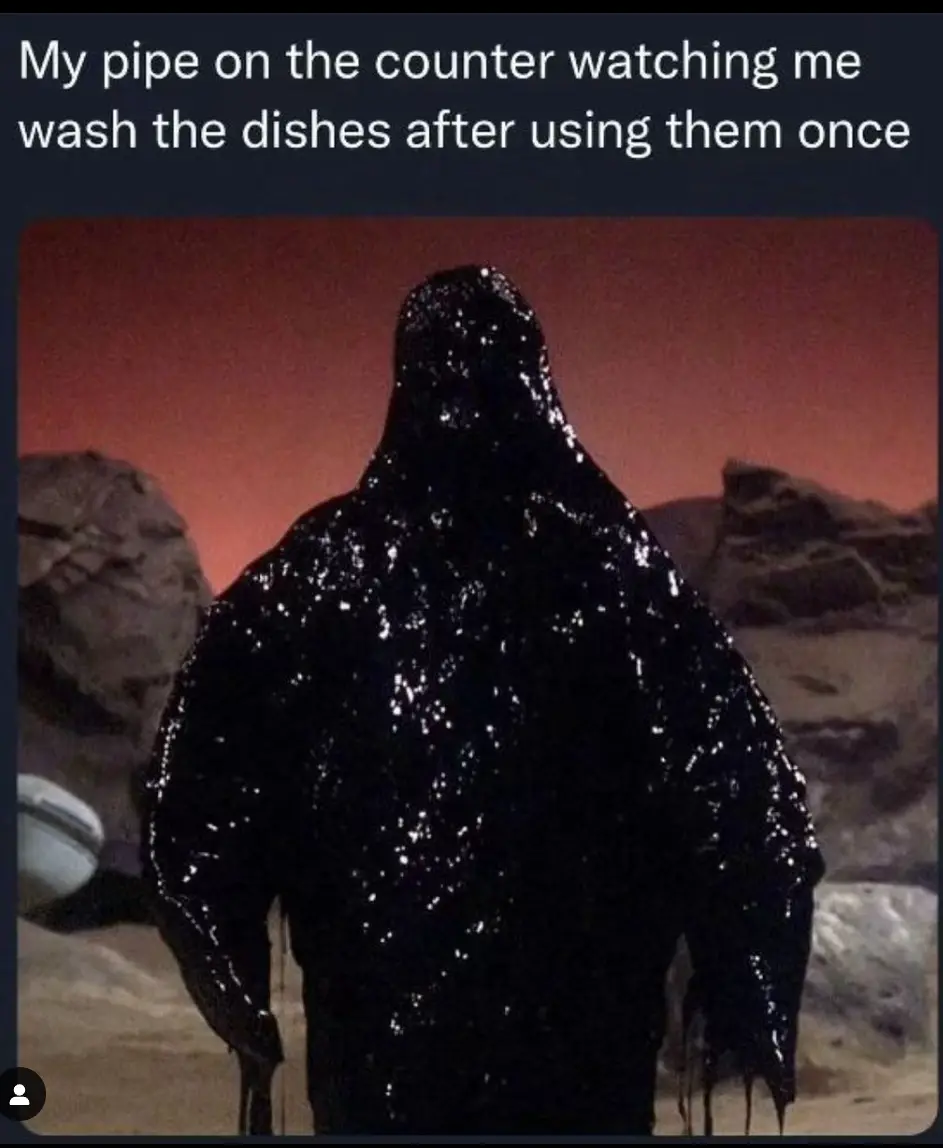 A dark, hooded figure made of black, shiny, dripping substance stands with its back to the viewer against a reddish sky and…