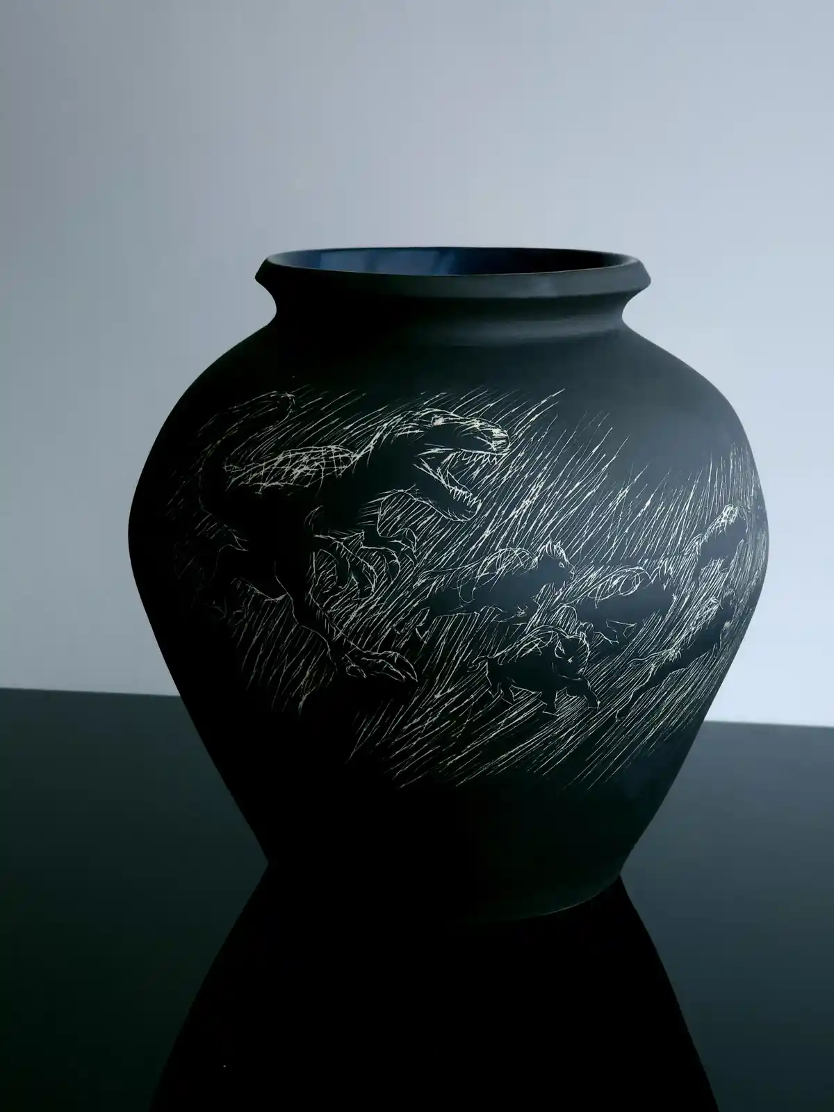 A black ceramic vase with a white etched scene of dinosaurs in chaotic motion, set against a plain gray background. The…