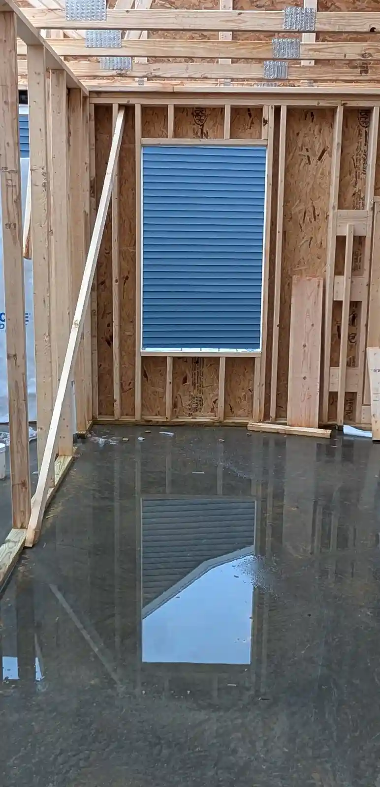 A construction site shows exposed wooden framing with a blue horizontal-louvered panel installed where a window or door…