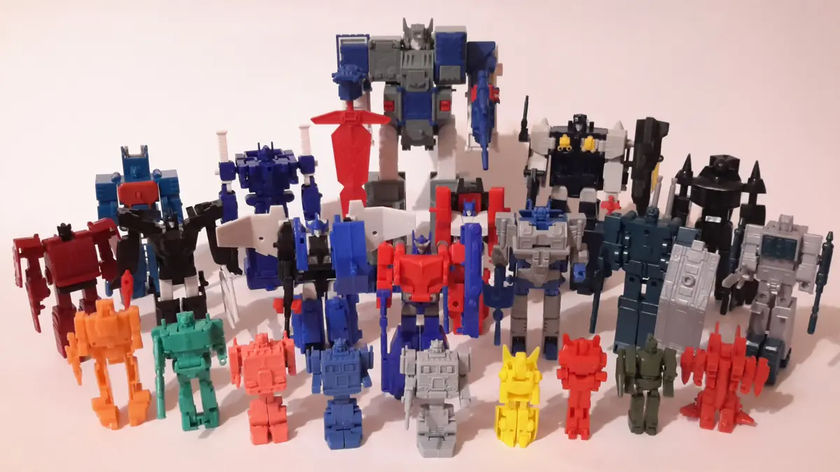 A large group of various Transformers action figures, mostly in robot mode, are arranged in rows on a plain white surface…