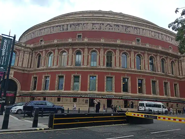 An exterior shot of the grand, red-brick Royal Albert Hall in London, featuring its iconic curved facade, ornate stone…