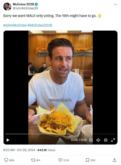 A man in a white t-shirt sits at a restaurant table holding a tray of loaded fries, looking slightly embarrassed or…