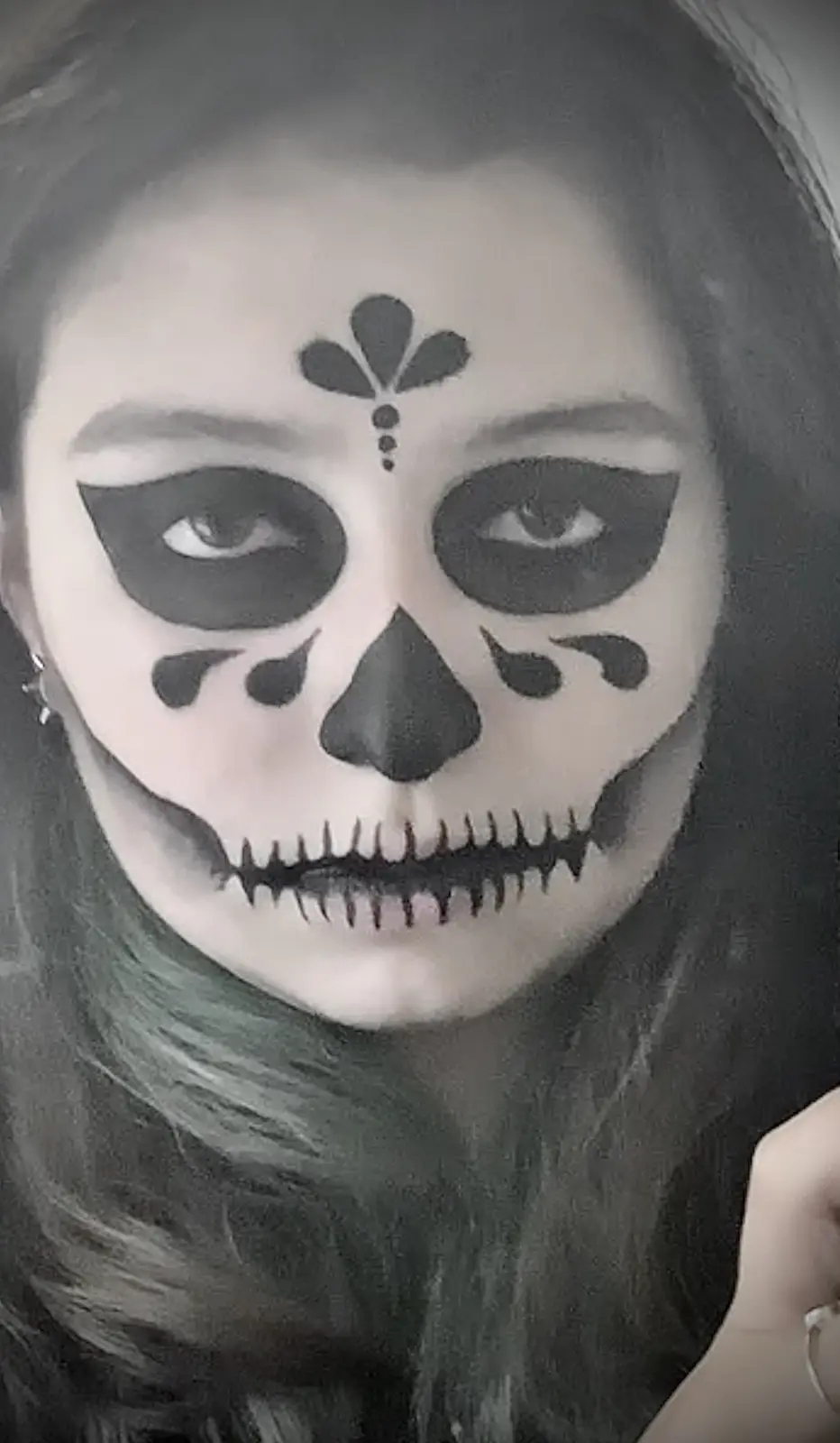 A person with long dark hair has intricate black and white sugar skull makeup on their face, featuring a floral design above…
