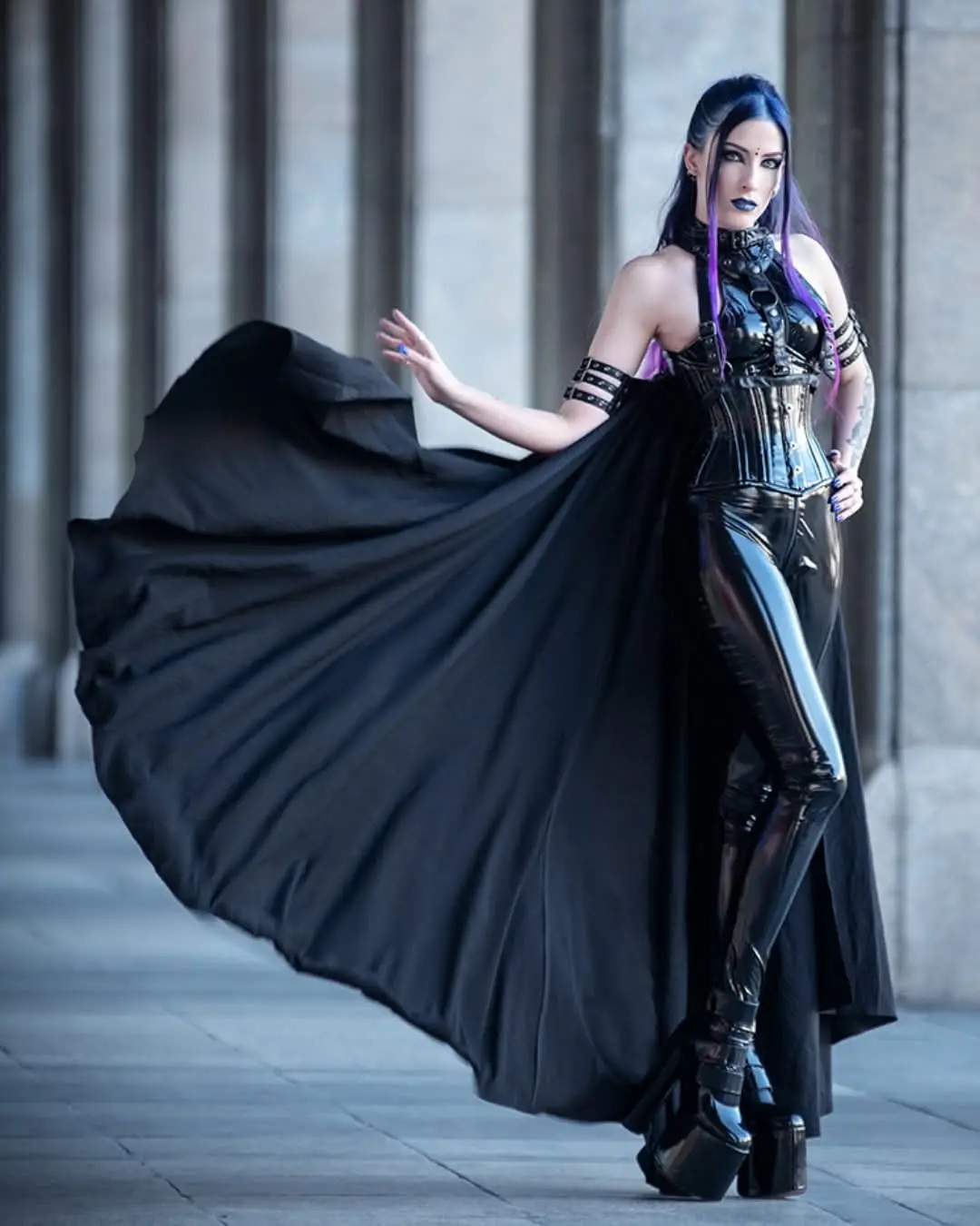 A woman with dark hair and purple streaks, wearing a shiny black corset, latex pants, and a flowing black cape, poses…