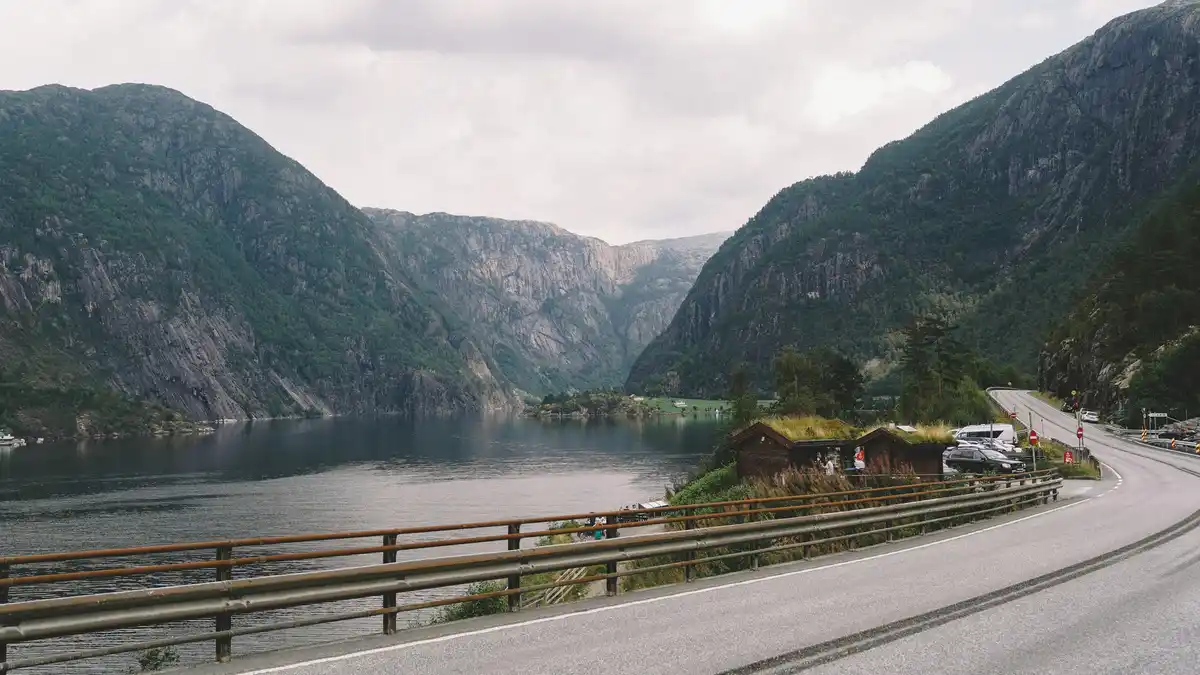 Winding road hugs the glassy fjord, mirrored mountains rising steeply from the water's edge, verdant forests a lush green…