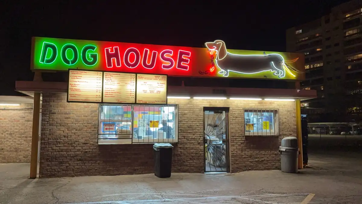 A nighttime photo of a brick restaurant with a large neon sign reading 'DOG HOUSE' in green and red letters, accompanied by…