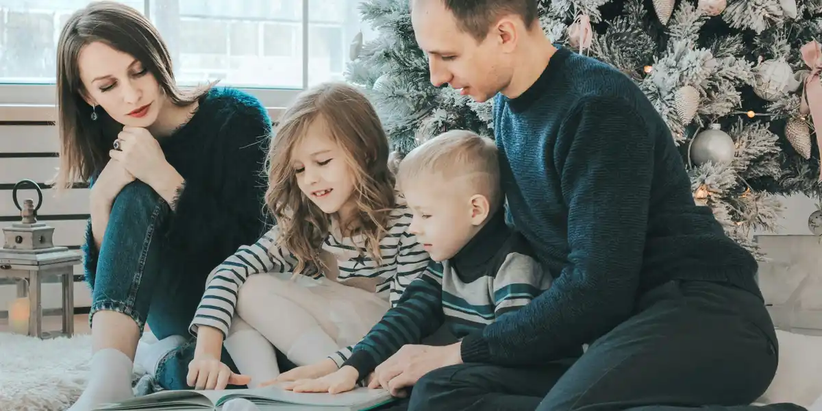 A family of four—mother, father, and two young children—are sitting on a white rug in front of a decorated Christmas tree…