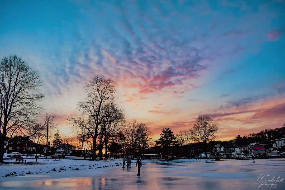 A serene winter scene shows people ice skating on a frozen pond under a vibrant sunset sky with pink, orange, and blue hues…