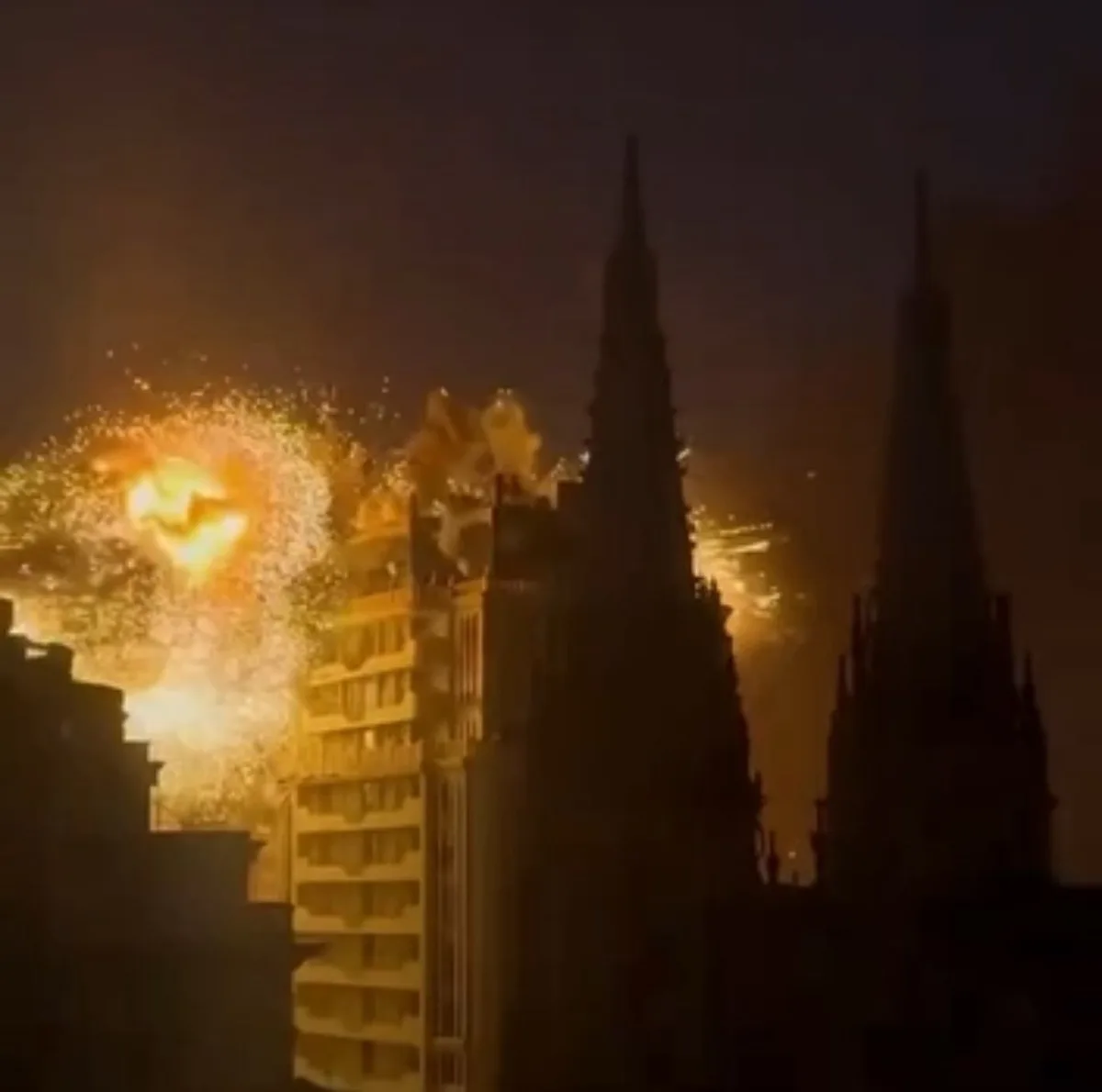 A nighttime cityscape featuring two tall, pointed spires silhouetted against a dark sky, with bright, golden fireworks…