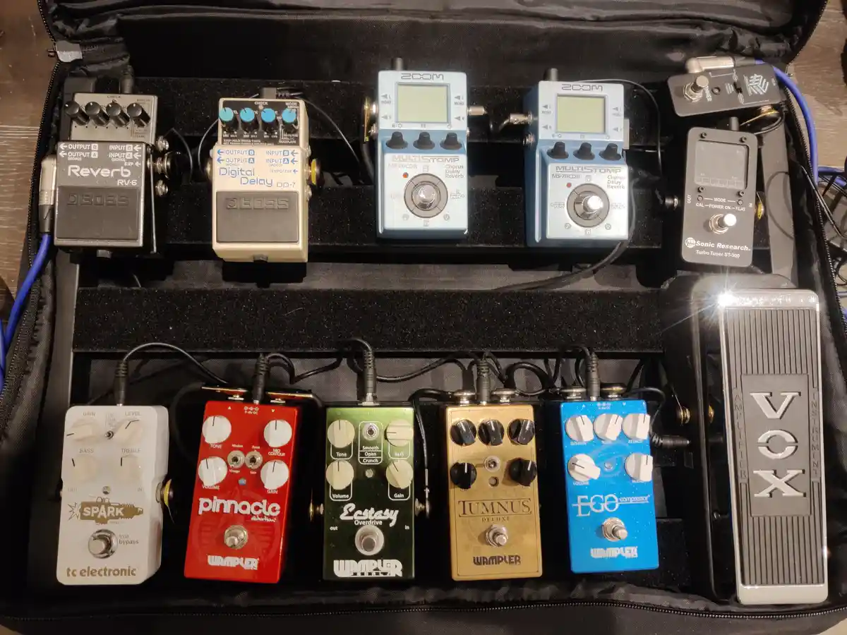 An overhead shot of a black pedalboard case filled with ten guitar effects pedals of various brands and colors, including…