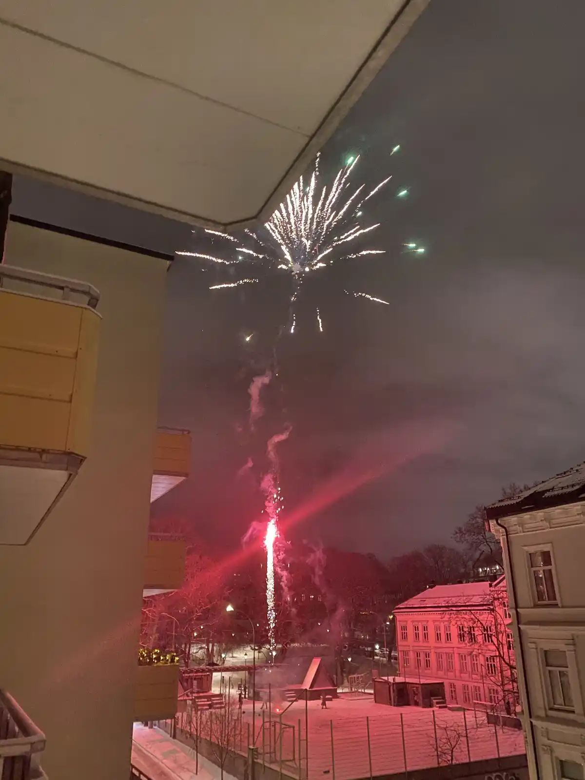 A nighttime photo taken from a balcony shows a large, bright fireworks explosion in the sky above a snowy urban area with…