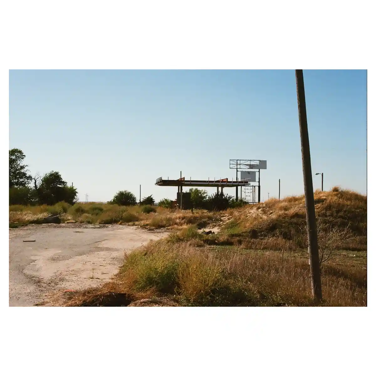 An abandoned, overgrown gas station with a rusted canopy and empty sign stands in a field of dry grass under a clear blue…