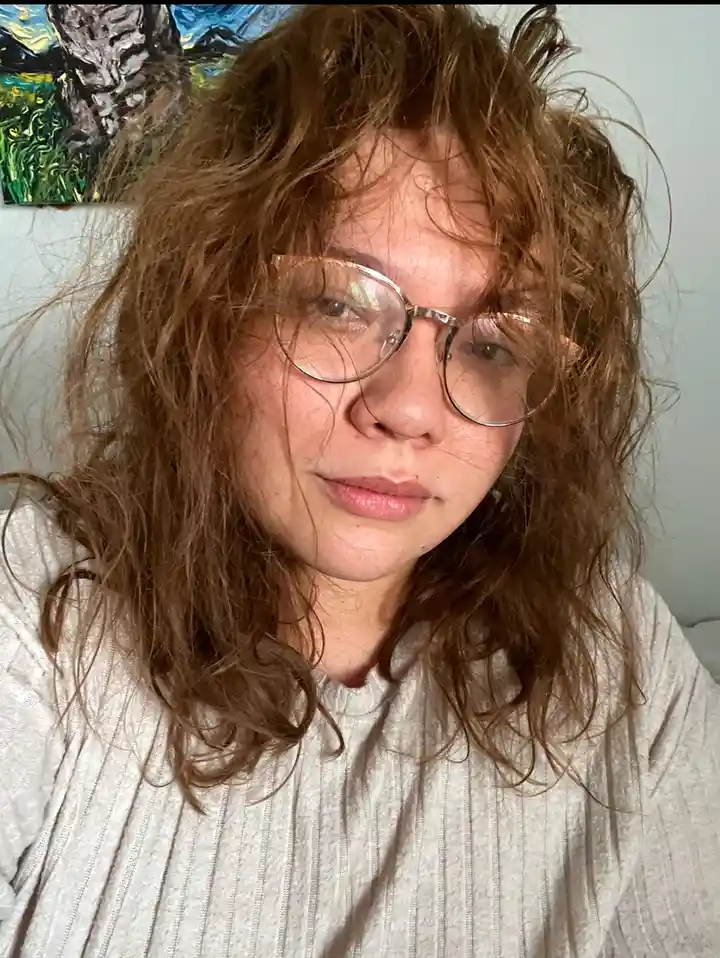A person with messy brown hair and round glasses looks directly at the camera with a tired, slightly annoyed expression…
