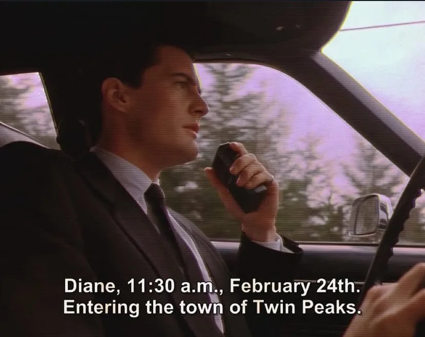 A man in a suit and tie sits in the driver's seat of a car, holding a walkie-talkie to his mouth as if reporting. The scene…