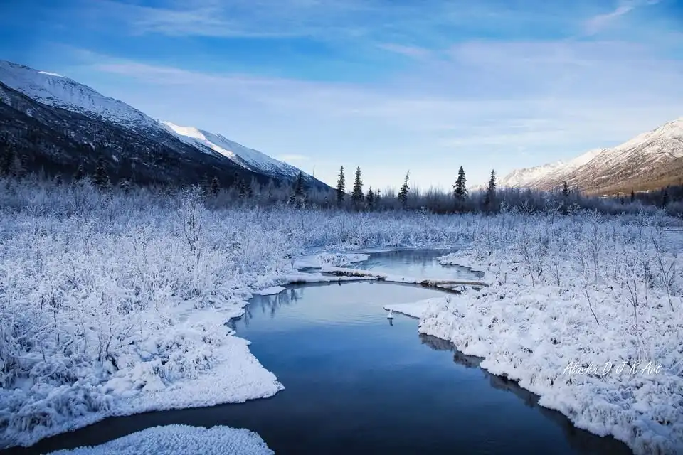 A serene winter landscape shows a winding river flowing through snow-covered bushes and trees, with snow-capped mountains in…