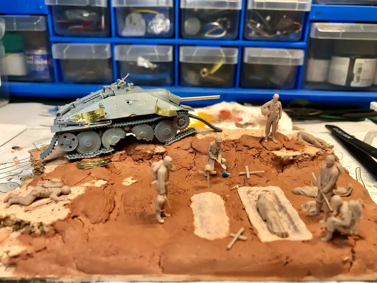 A diorama of a model tank and soldiers on a reddish-brown clay battlefield with several figures lying down as if dead, some…