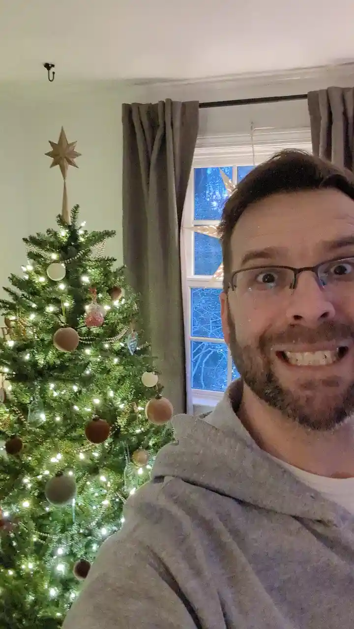 A man with glasses and a beard makes a wide-eyed, exaggeratedly shocked expression while taking a selfie in front of a…