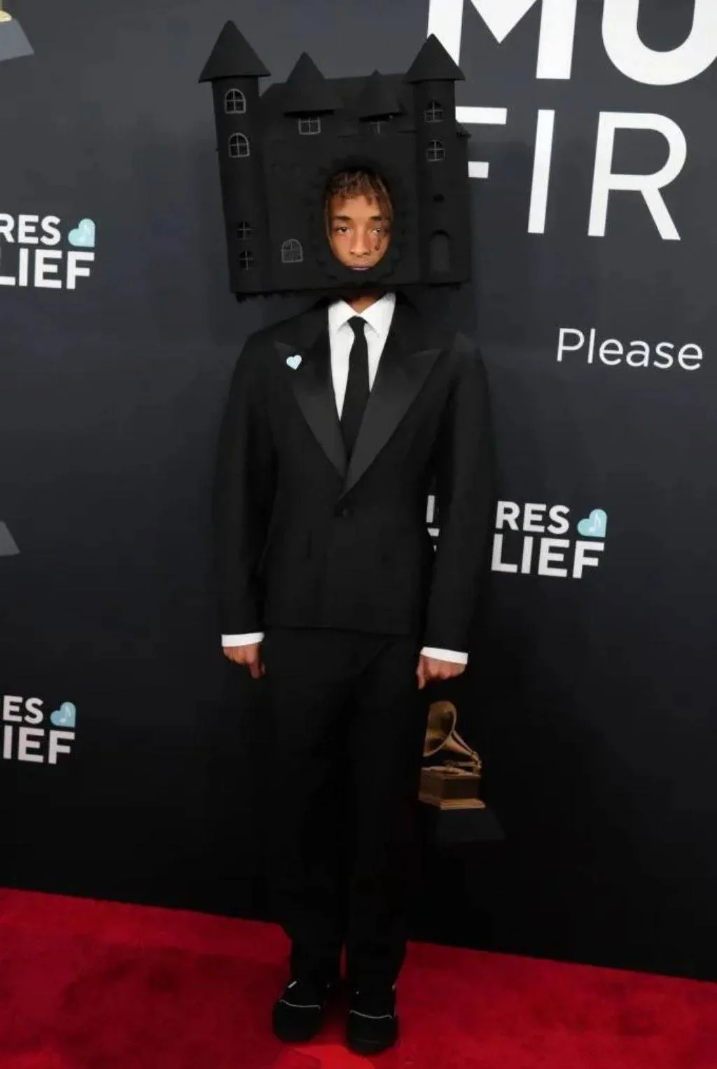 A man in a black tuxedo stands on a red carpet holding a Grammy award, wearing a large black cardboard castle on his head…