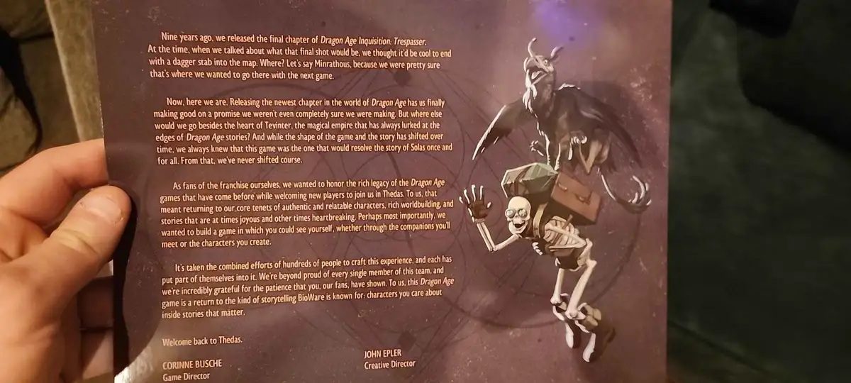 A hand holds up a printed page from a Dragon Age game, showing a paragraph of text from a game developer's statement. The…