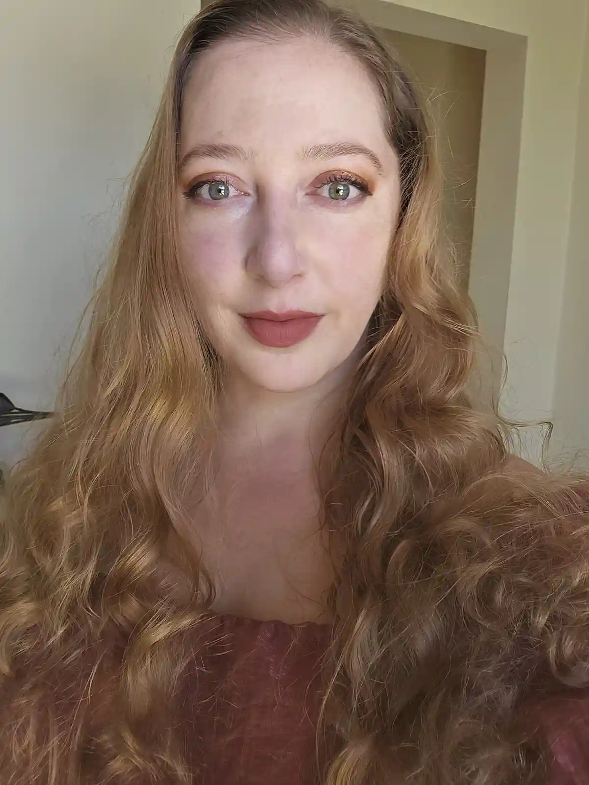 A woman with long, wavy blonde hair and makeup looks directly at the camera with a neutral expression. She wears a…