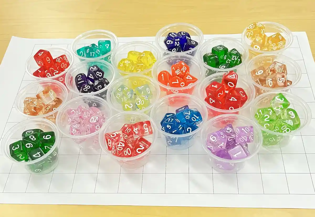 An overhead shot shows numerous small, clear plastic cups arranged on a white gridded sheet, each filled with colorful…