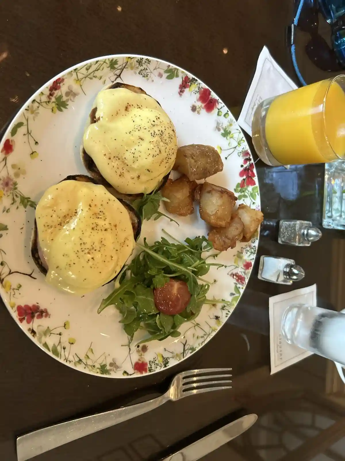 A top-down photo of a plated breakfast featuring two eggs benedict with hollandaise sauce, roasted potatoes, and a small…