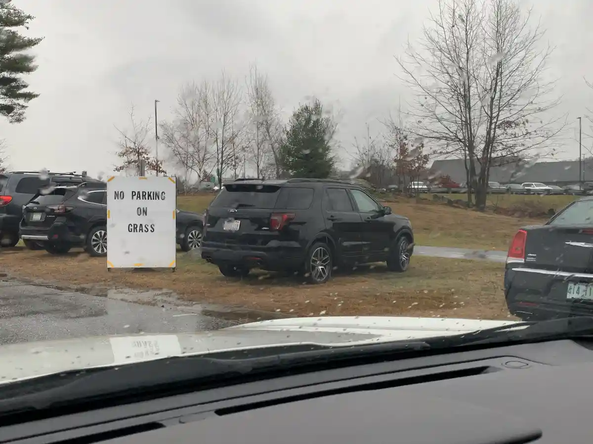 A photo taken from inside a car shows a black SUV parked on grass next to a white sign that reads 'NO PARKING ON GRASS'. The…