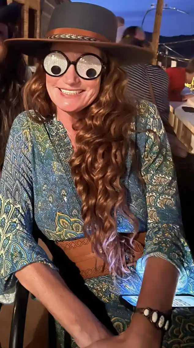 A woman with long curly hair, wearing a wide-brimmed hat and a patterned blue-green top, smiles while seated. Her face is…
