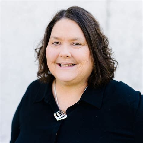A smiling woman with shoulder-length brown hair wearing a black collared shirt and a silver pendant necklace stands against…