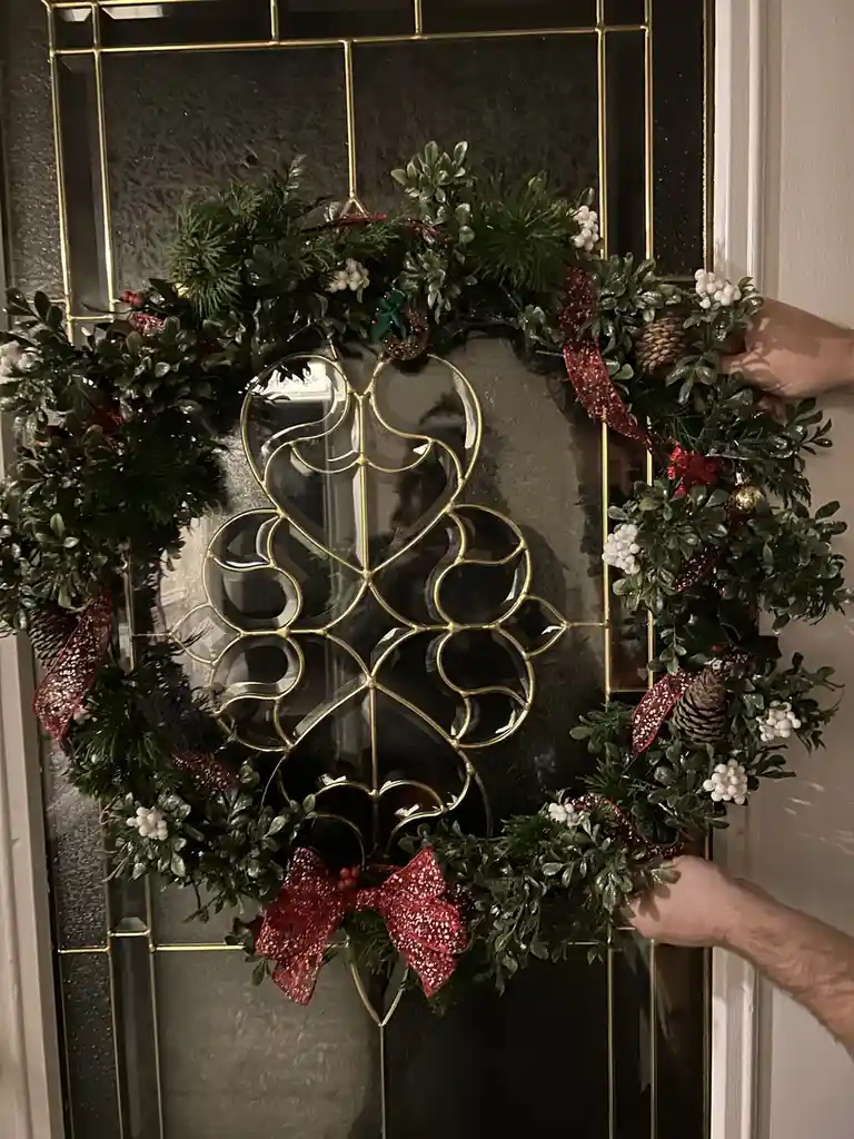 Two hands are shown hanging a festive Christmas wreath adorned with greenery, pinecones, white berries, and red glittery…