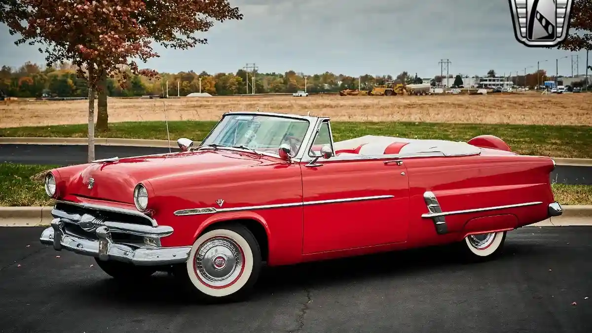 a shiny red 1955 ford sedan convertible with white wall tires and chrome trim is parked on asphalt, with a tree and open…